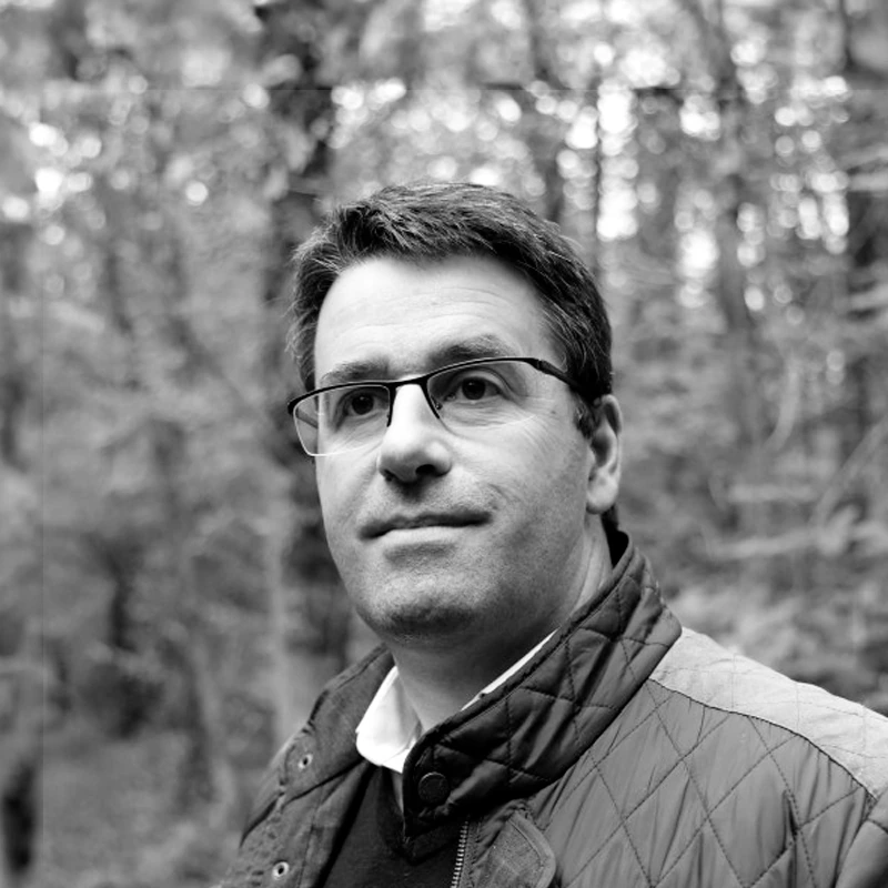 Portrait en noir et blanc de Sebastien, expert forestier, en veste matelassee devant une foret